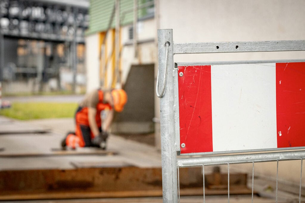 Hinter einer Baustellen-Absperrung sieht man im Hintergrund einen Servicetechniker in ESSMANN-Arbeitskleidung knieend arbeiten. Hinter ihm sieht man eine Fabrik als Kulisse.