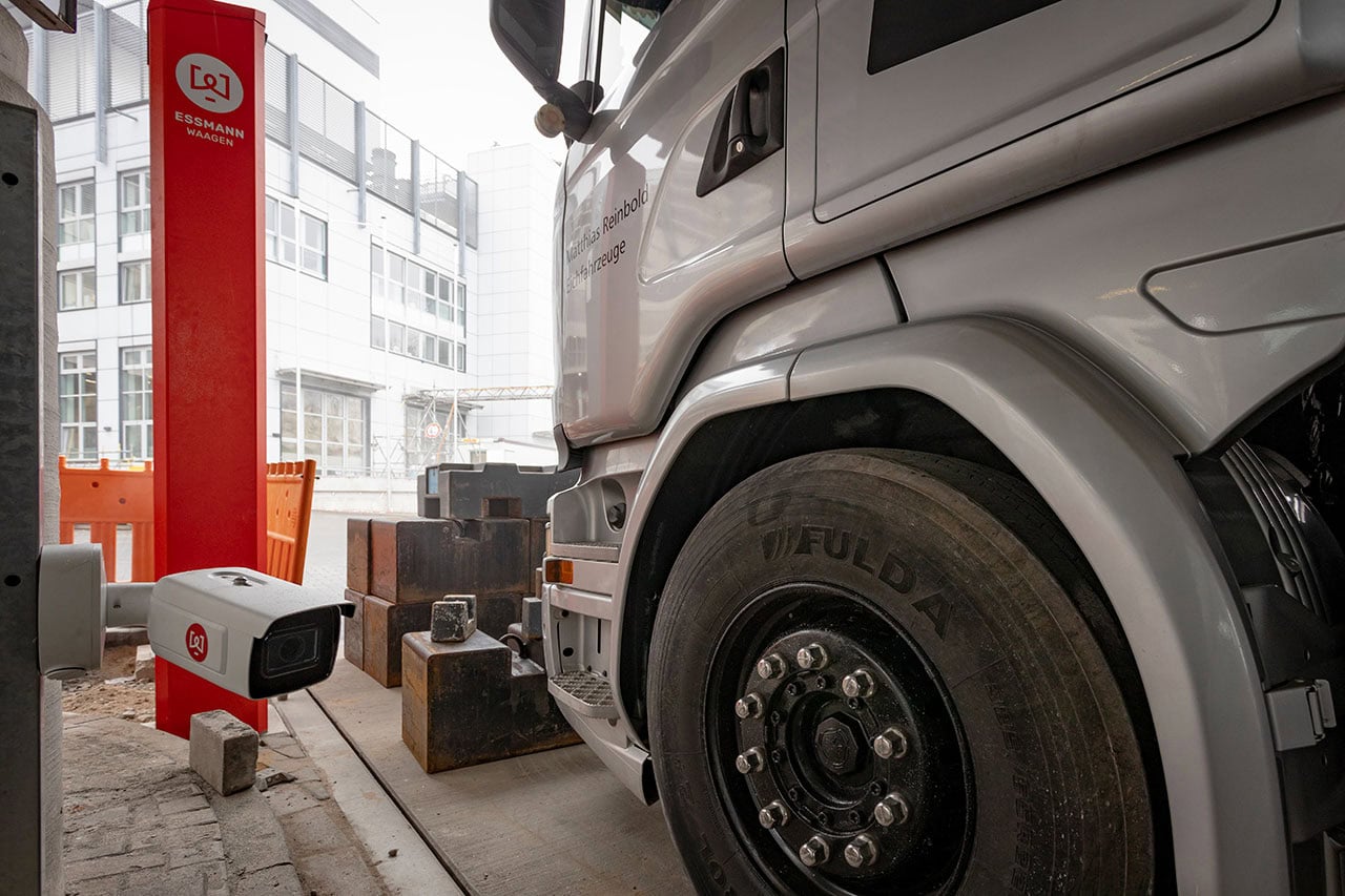 Gewichte und Eich-LKW auf Fahrzeugwaage neben roter Selbstbedinesäule und Kamera zur Kennzeichenerkennung.
