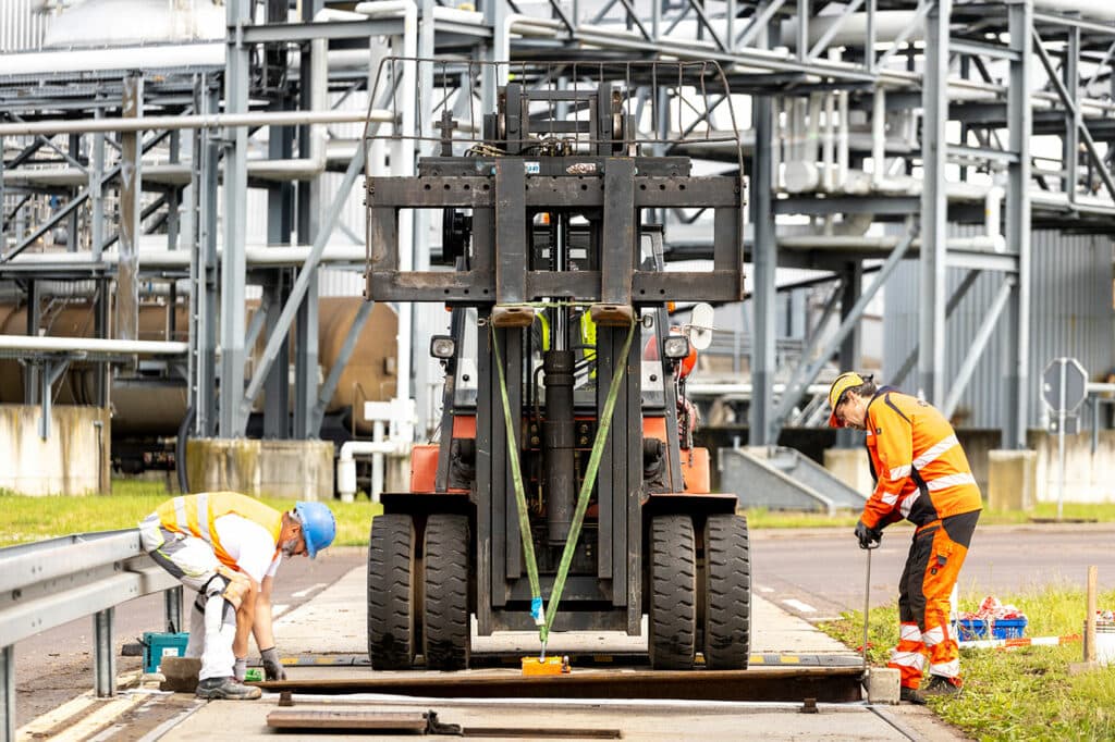Gabelstapler hebt mit Magnet Metallabdeckung auf die LKW-Waage.Ein Werksarbeiter und unser Servicetechniker führen die Platte ein.