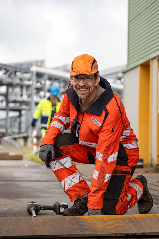 Ein freundlich lächelnder Servicetechniker in oranger Arbeitskleidung und Anstoßkappe kniert auf einer Fahrzeugwaage und schaut lächelnd in die Kamera.