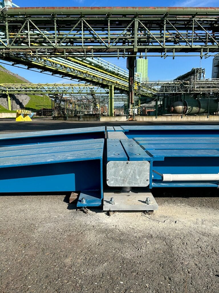 Industriële installatie met blauwe HE-TS weegbrug vanaf de zijkant onder leidingwerk