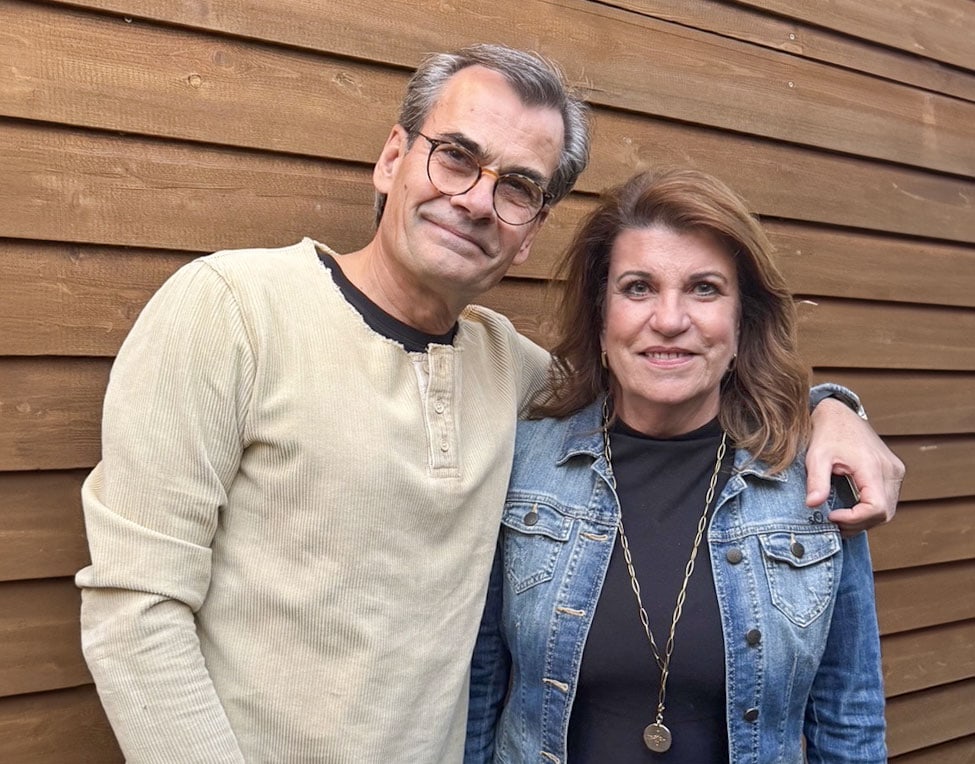 Holger Thiel und Barbara Visser-Thiel vor einer hölzernen Wand. Holger Thiel links trägt ein beiges Langarmshirt, Barbara Visser-Thiel rechts eine Jeansjacke und Halskette. Der Arm von Holger liegt auf Barbaras Schultern.